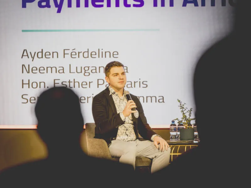 Ayden Férdeline holding a microphone, seated on a chair on stage, giving a presentation. Behind him, part of his slide is visible on a large screen. In the foreground, the silhouettes of audience members are visible between the camera and the speaker.