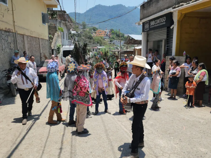 Banner image of a lively street scene in rural Mexico, with people in traditional costumes and masks dancing. A young man stands in the center playing a guitar, surrounded by colorful celebration.