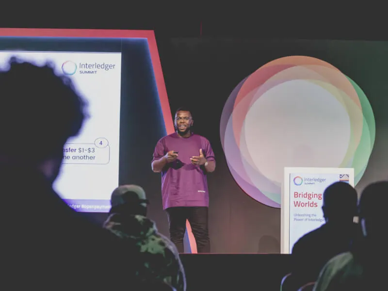 Uchi Uchebeke stands on stage giving a presentation at an Interledger Summit, with audience silhouettes visible in the foreground between him and the camera.
