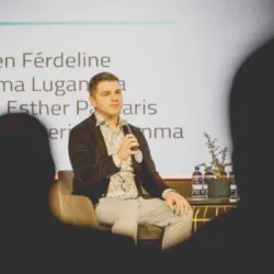 Man presenting on stage with microphone, audience silhouettes in foreground.