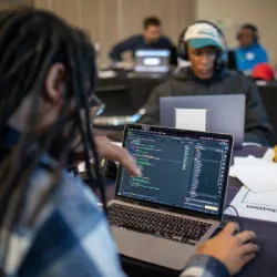 Close-up of a laptop screen displaying code, with a blurred user in the foreground and other hackathon participants working on laptops in the background.